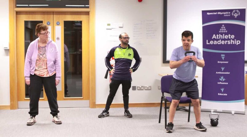 Two people watch as a fitness leader demonstrates a fitness exercise using a kettlebell.
