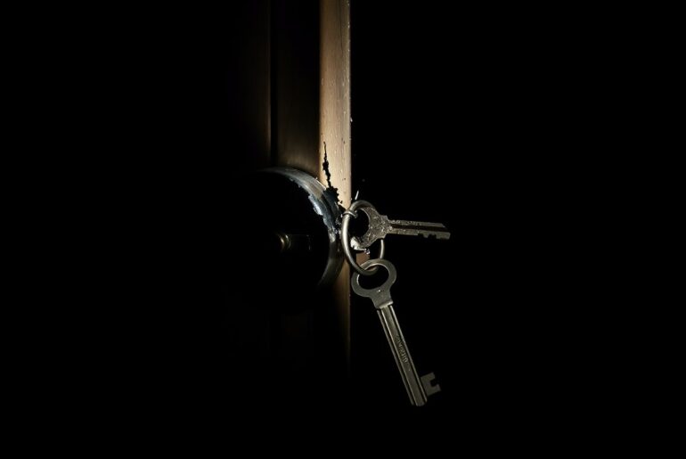 An extreme close-up photograph of a broken lock or key, lit by a harsh, direct camera flash against a pitch-black background, creating a stark, gritty, investigative aesthetic.