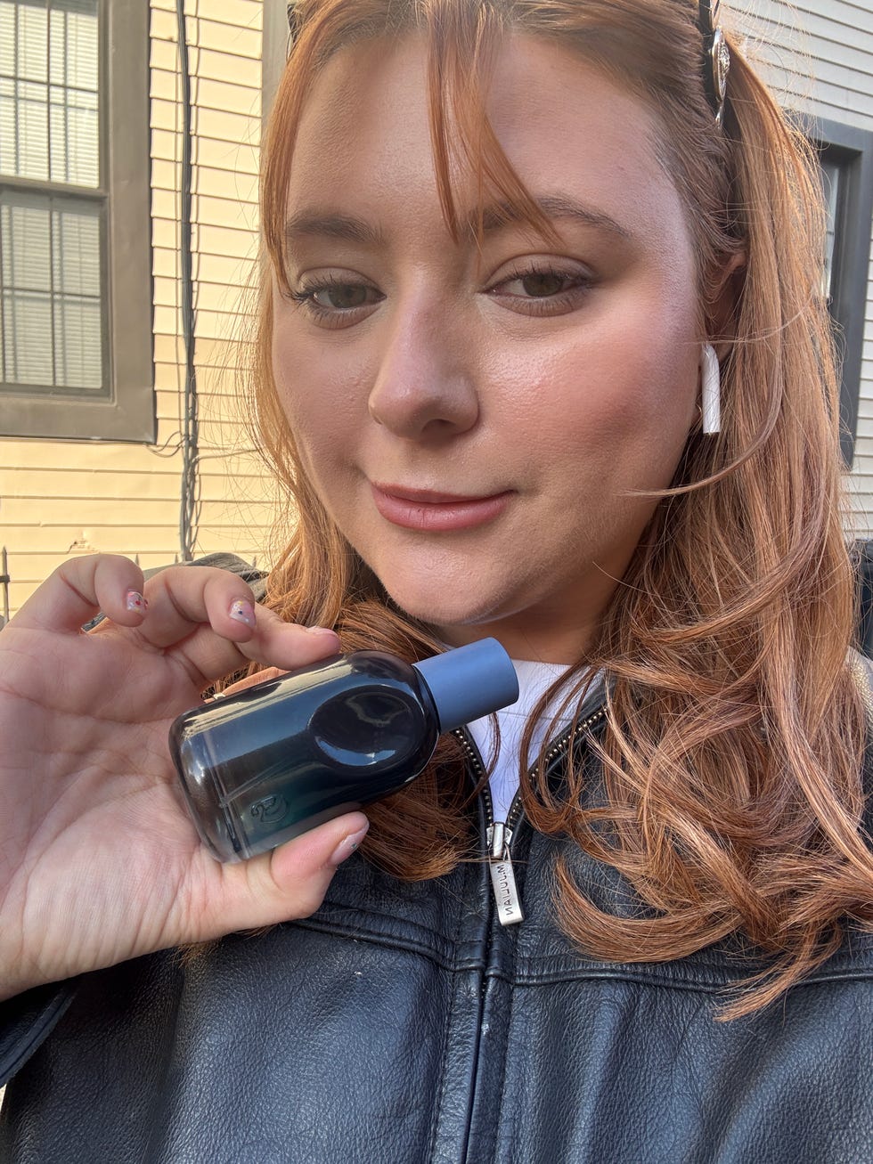 A person holding a small bottle in front of their face, with residential architecture in the background.