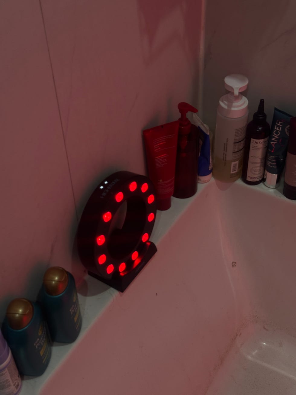 Illuminated device and various hair and skincare products arranged on the edge of a bathtub.