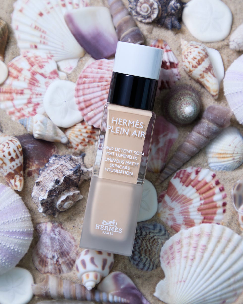 Makeup foundation bottle among seashells on sand.