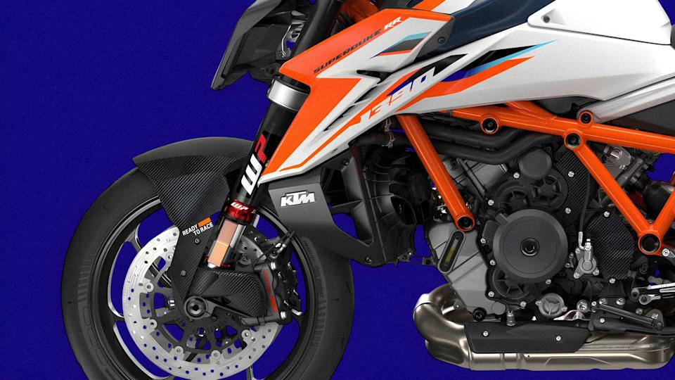 Close-up of the front wheel, suspension, and engine frame of an orange and white KTM Super Duke RR motorcycle against a blue background.