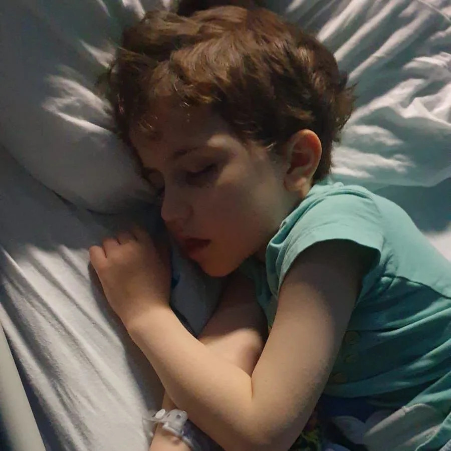 A young child lying asleep on a bed, with a bandage on one hand and holding a soft toy.