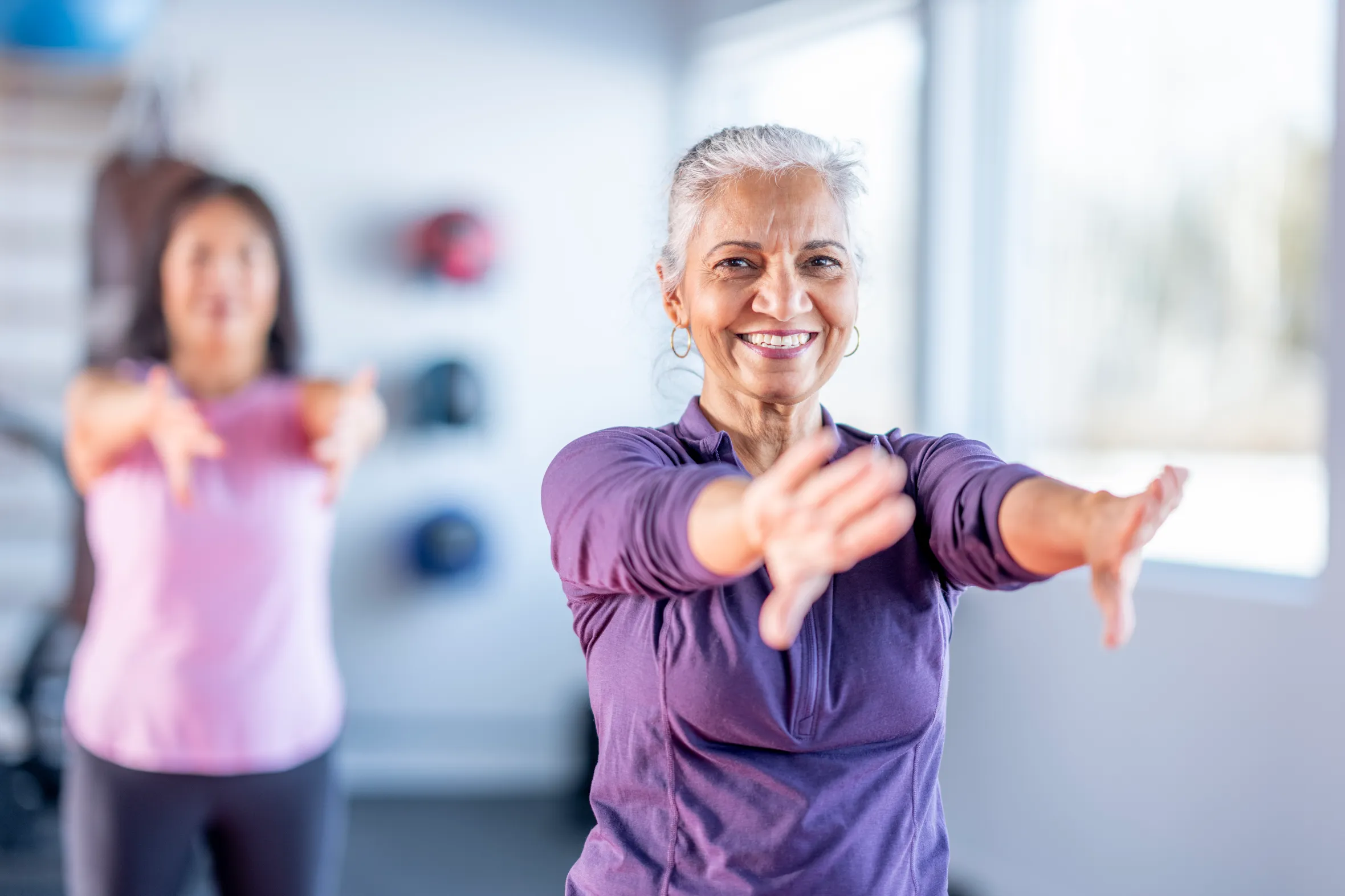 A smiling senior woman in a purple top leads an arm-stretching fitness class.