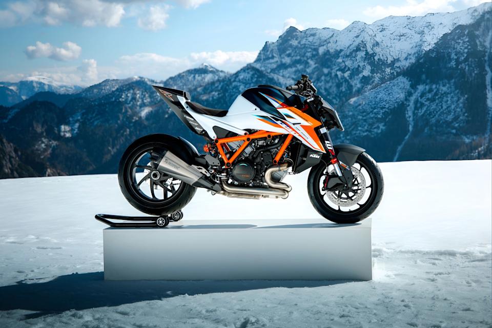 White, black, and orange KTM motorcycle on a stand in a snowy mountain landscape.
