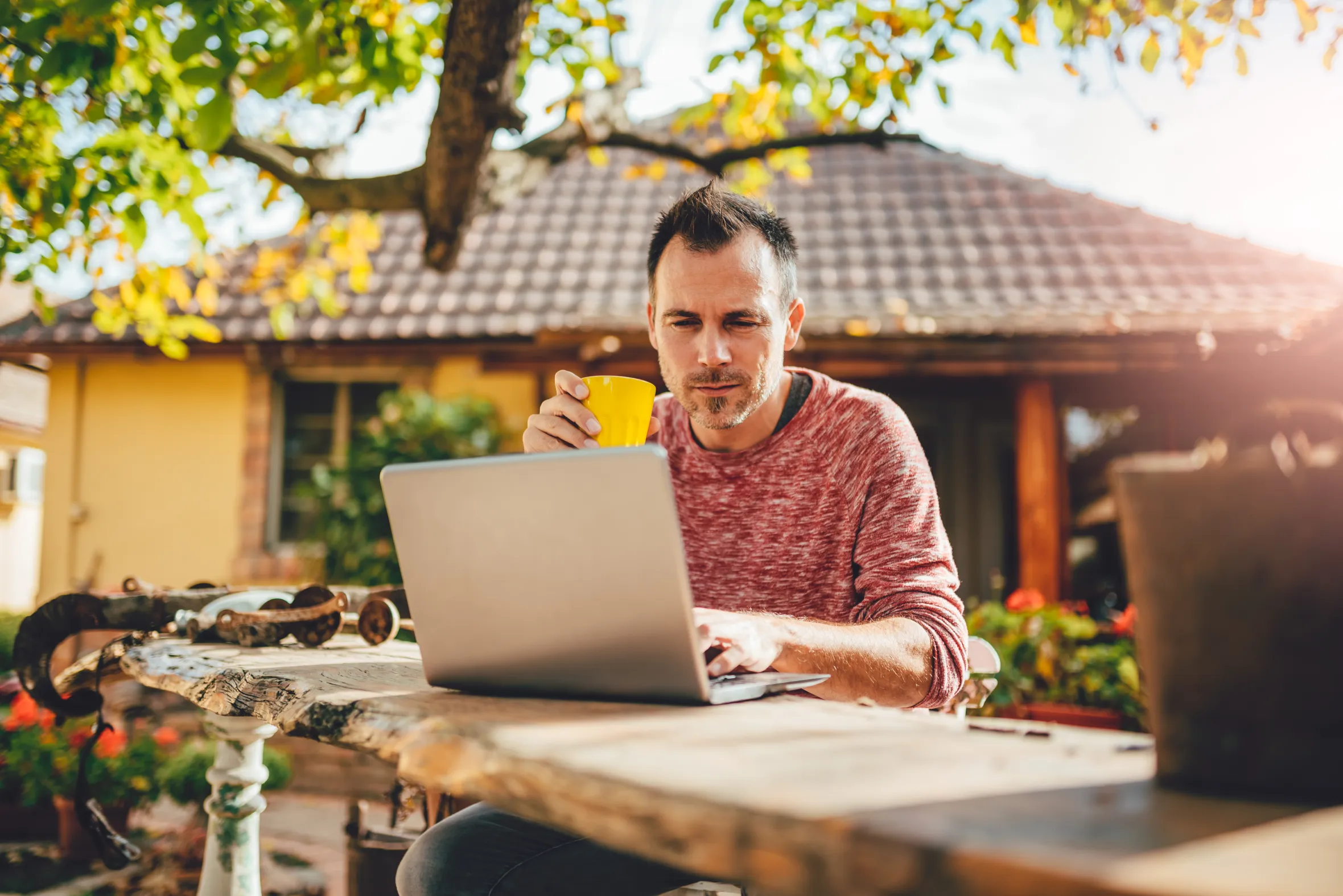 A man sitting at a wooden table in a backyard patio, drinking coffee and using a laptop.
