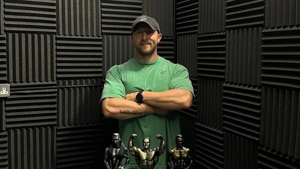 Sion standing with his arms crossed in front of him. He smiles at the camera in front of a black wall. Sion wears a green sports t-shirt and black cap. In front of him are three body building trophies. 