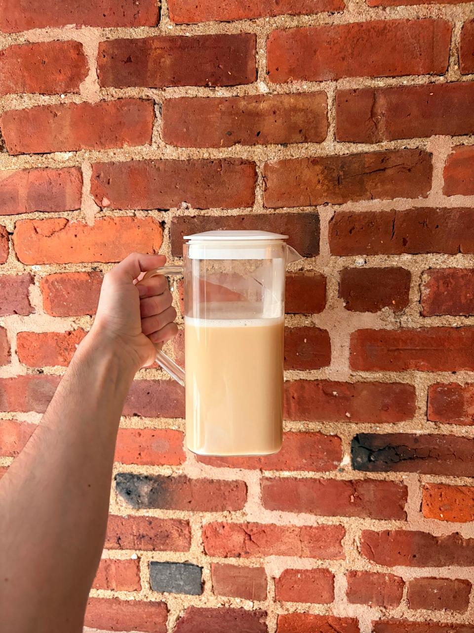 A hand holding a clear pitcher filled with a light-colored beverage against a brick wall.