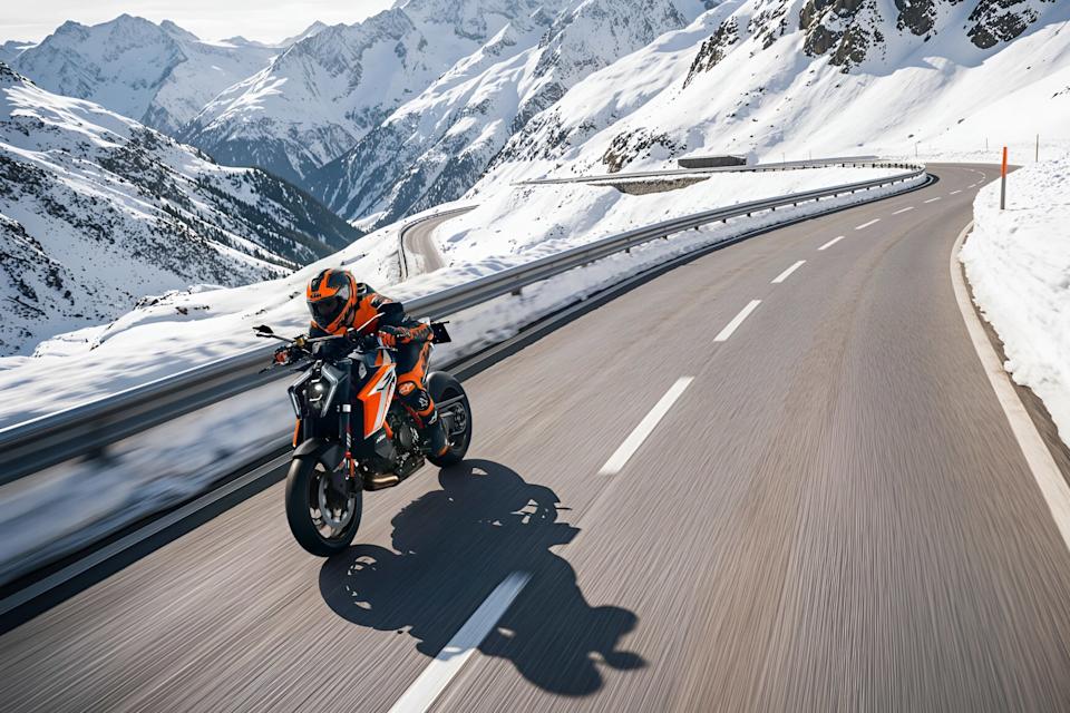 Motorcyclist in orange gear riding a black and orange KTM bike on a winding mountain road with snow-covered peaks.