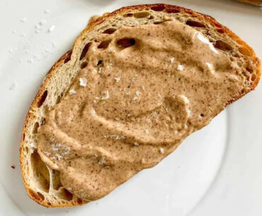 Homemade almond butter spread on a slice of artisanal bread, on a white plate with a silver knife resting on the plate.