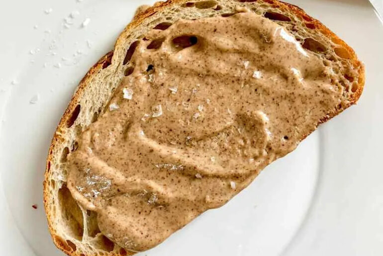 Homemade almond butter spread on a slice of artisanal bread, on a white plate with a silver knife resting on the plate.