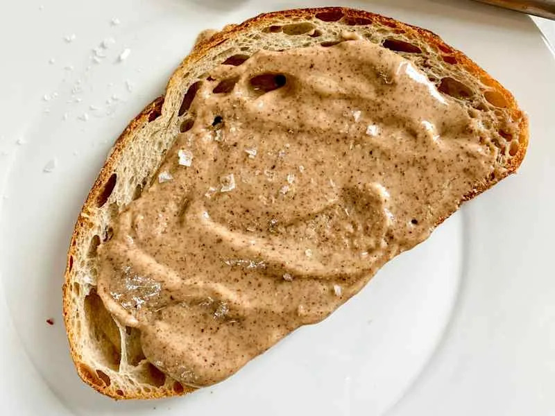 Homemade almond butter spread on a slice of artisanal bread, on a white plate with a silver knife resting on the plate.