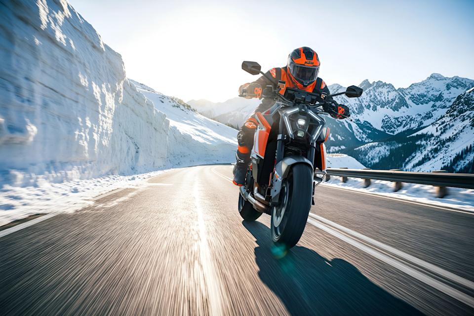 Motorcyclist in orange and black gear riding a KTM motorcycle on a snowy mountain road at sunrise.