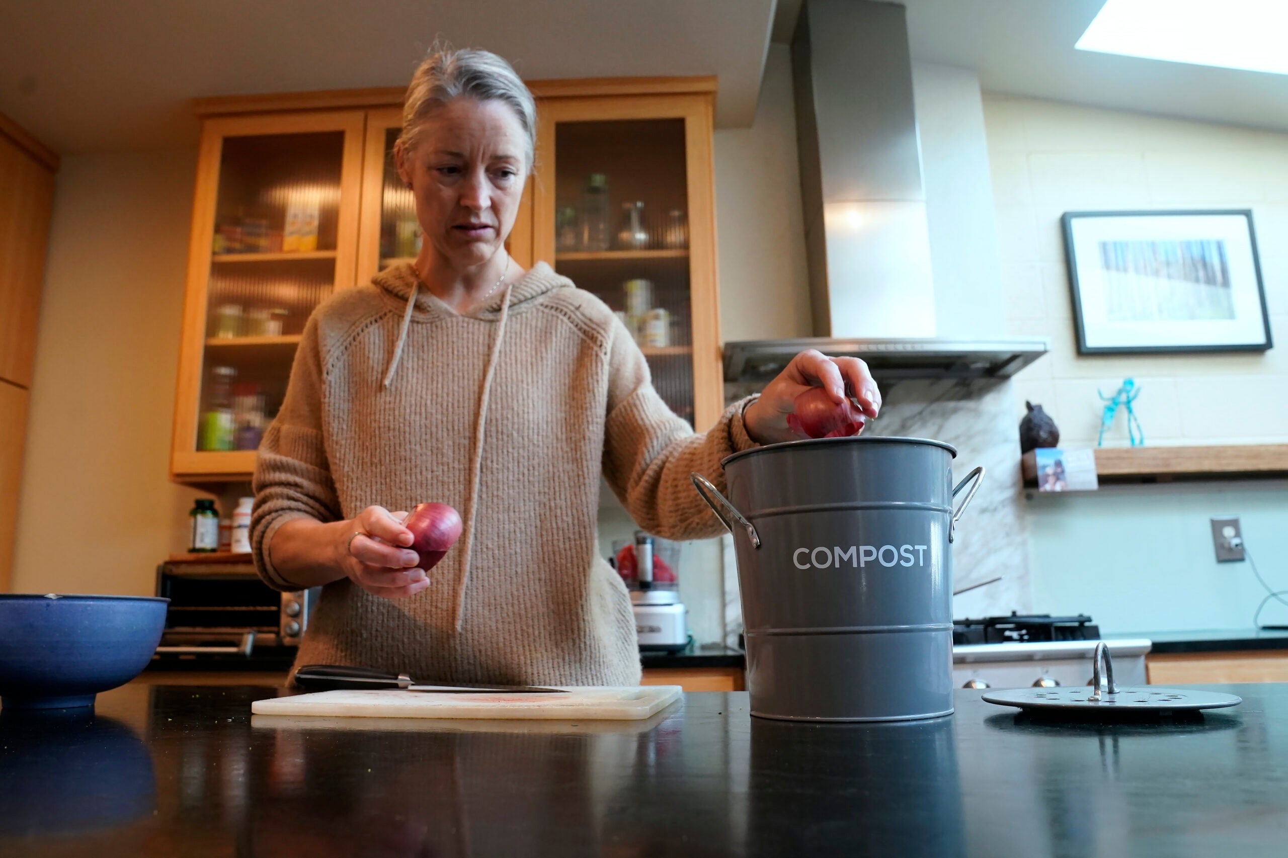 A woman in a kitchen places food scraps into a metal compost bin labeled COMPOST while holding an onion.