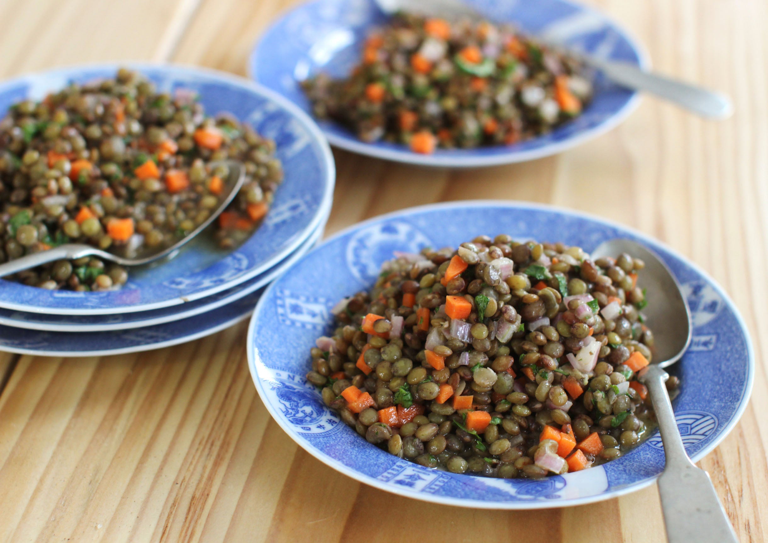 Three blue plates each contain a serving of lentil salad with diced carrots and ham on a wooden table, with spoons beside the plates.