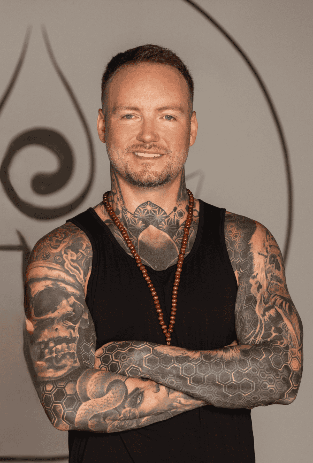 Portrait of a smiling man with short hair, tattoos on neck and chest, wearing a beaded necklace.