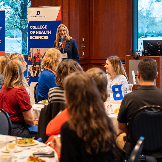Amanda Leightner speaks into a microphone as tables full of students and community members listen.