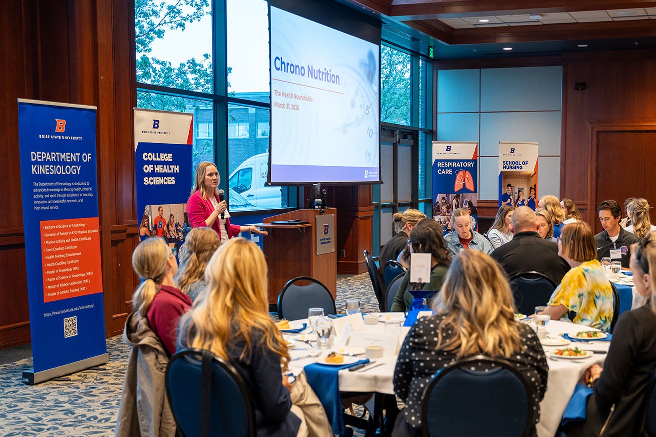 People sit at tables listening to Sara Weelborg speaking into a microphone.