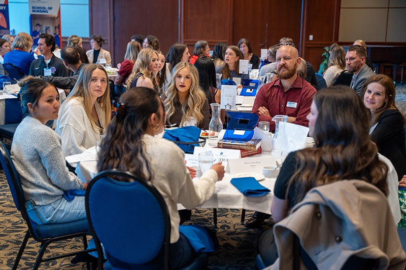 In a crowded room, people sitting at one table look at a student while listening to her.
