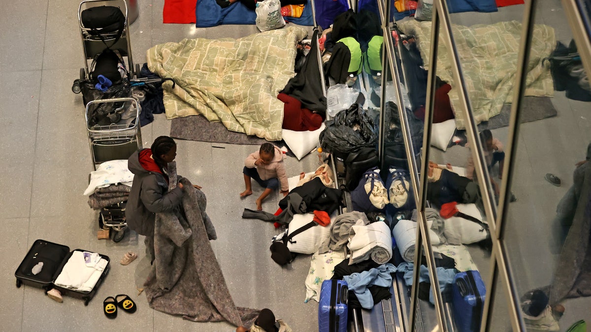 Migrant mother folding blanket next to three children sleeping on terminal floor
