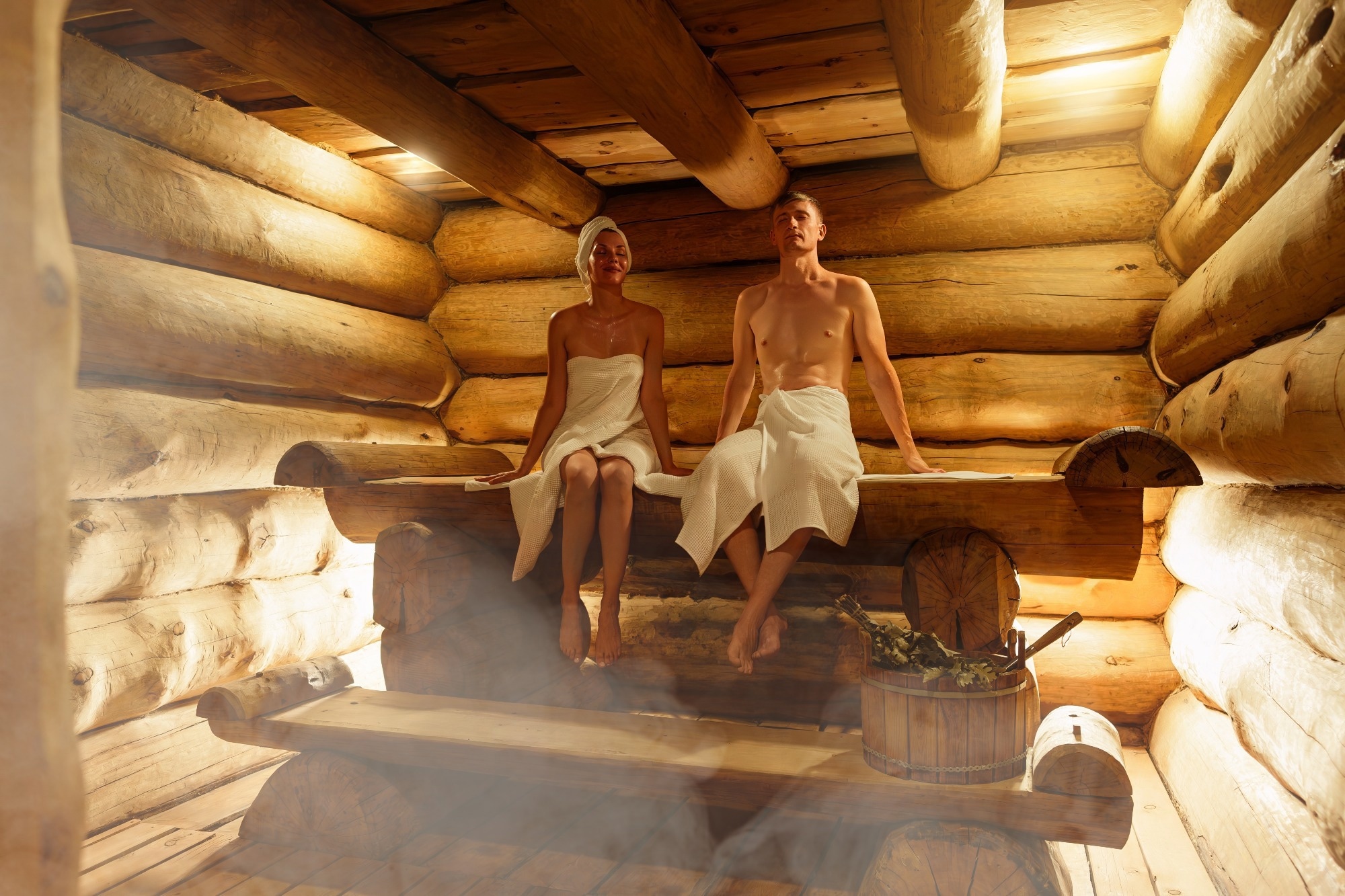 Young couple relaxing and sweating in hot sauna wrapped in towel. Woman and man In Sauna. Interior of Finnish sauna, classic wooden steam room with hot steam. Russian bathroom. Relax in hot Bathhouse.