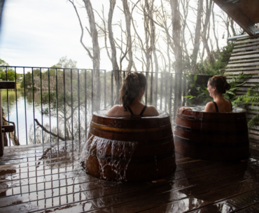 Two individuals are relaxing in wooden hot tubs on a deck beside a serene lake of the Metung Hot Springs. The tubs are steaming, and the area is surrounded by trees and wooden fencing. The scene suggests a peaceful and natural retreat.