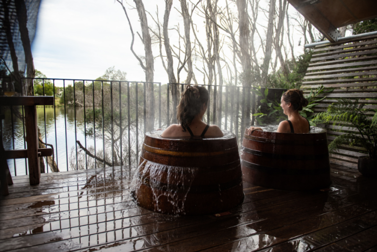 Two individuals are relaxing in wooden hot tubs on a deck beside a serene lake of the Metung Hot Springs. The tubs are steaming, and the area is surrounded by trees and wooden fencing. The scene suggests a peaceful and natural retreat.