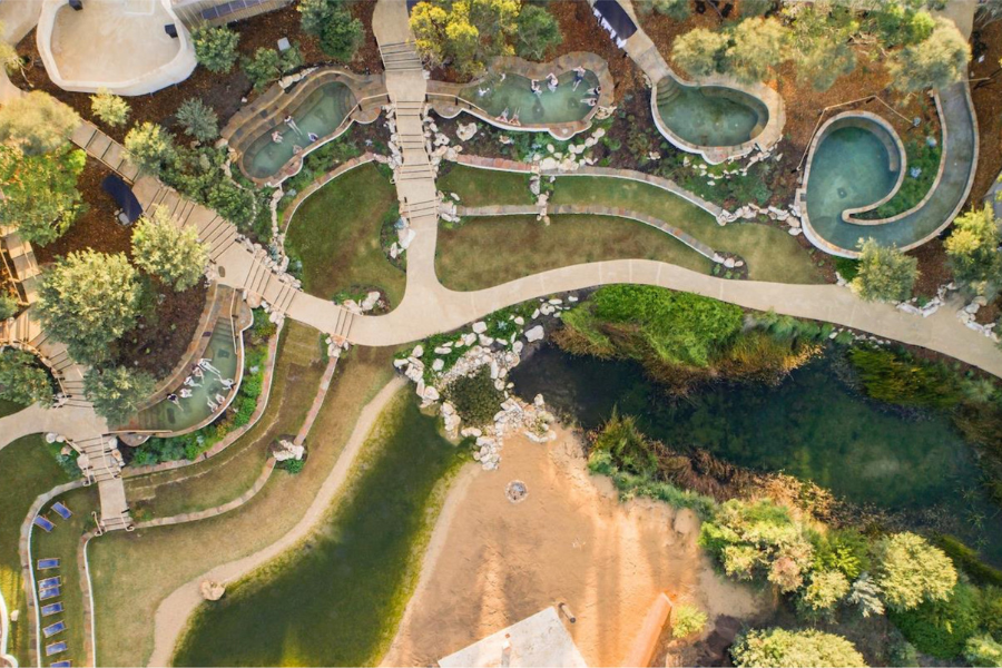This is an aerial view of a lush, landscaped area featuring winding pathways, small pools, and rock formations surrounded by greenery. The scene includes a mix of pathways, vegetation, and hot springs. 