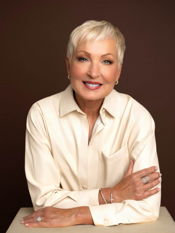 EIM Trainer Cindy Pounds, a smiling older woman with short blonde hair, wears a cream-colored button-up shirt, silver rings, and a bracelet as she sits with her arms folded on a table against a plain brown background.