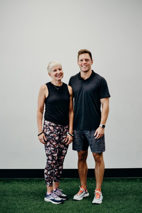 EIM Trainer Cindy Pounds stands with her son, side by side in athletic wear, smiling in front of a plain white wall on a green indoor surface.