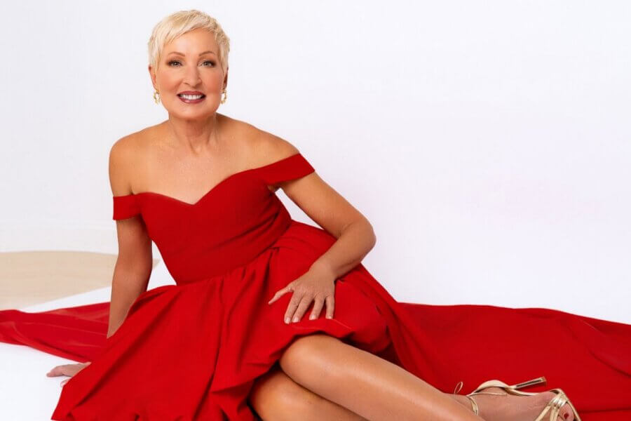 EIM Trainer Cindy Pounds, with short blonde hair, sits on the floor in an off-the-shoulder red dress and gold heels, smiling at the camera against a plain white background.