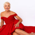 A woman with short blonde hair wearing an off-the-shoulder red dress sits on the floor, smiling at the camera against a plain white background, radiating confidence like an eim personal training success story.