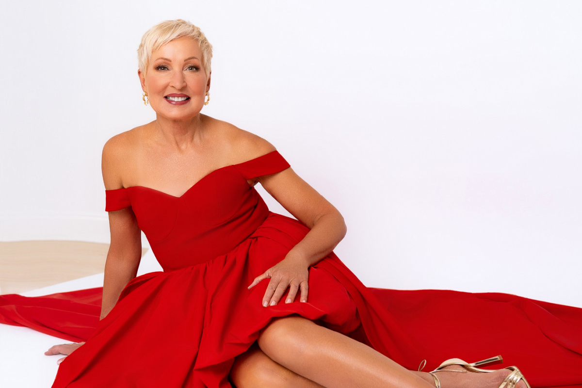 A woman with short blonde hair wearing an off-the-shoulder red dress sits on the floor, smiling at the camera against a plain white background, radiating confidence like an eim personal training success story.