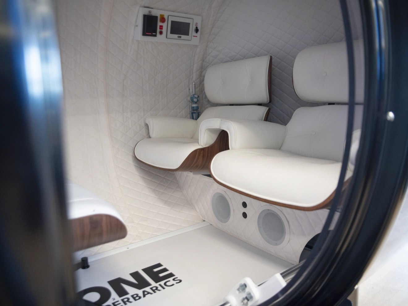 Interior of a hyperbaric chamber with white padded seats and control panel on the wall.