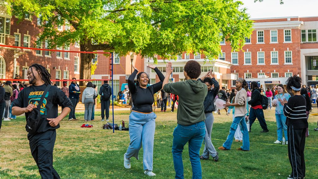 Wide shot of the quad with lots of students playing games like volleyball. In the center, two people give each other a high five.