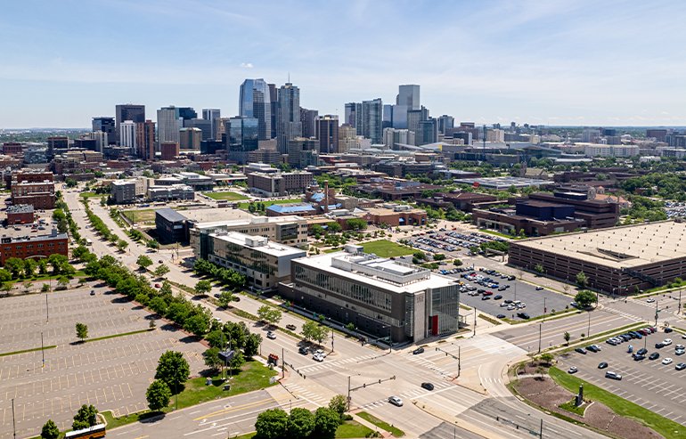 Aerial view of downtown Denver with campus buildings, streets, and parking lots in the foreground under a clear sky.