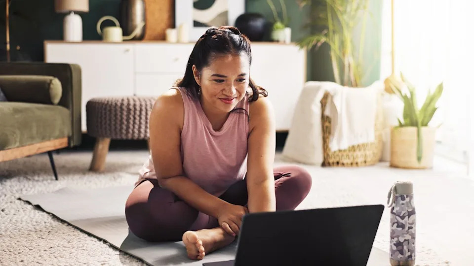  Woman sits cross-legged on yoga mat with open laptop and water bottle on floor in front of her. 