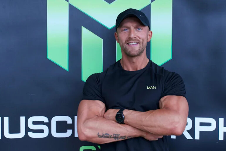 Sion standing with his arms crossed in front of a black branded gym backdrop. He wears a black t-shirt and a black cap. He smiles while looking at the camera.
