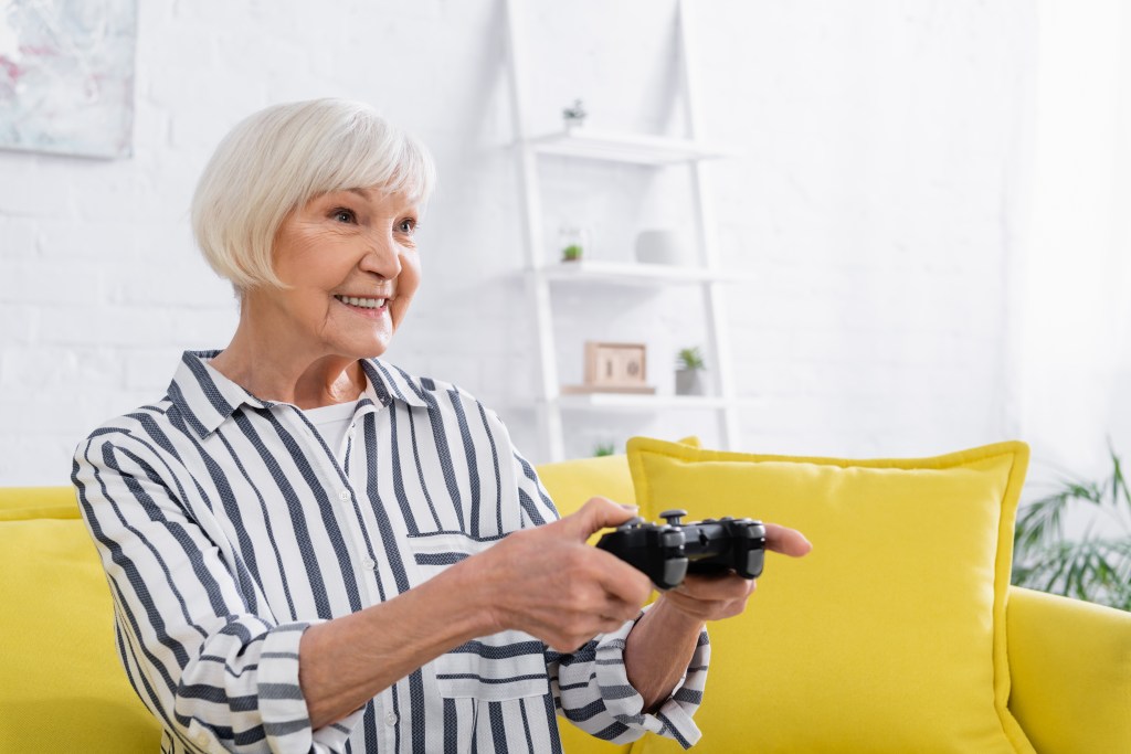 Elderly woman in striped shirt sitting on a yellow sofa and smiling while playing a video game.