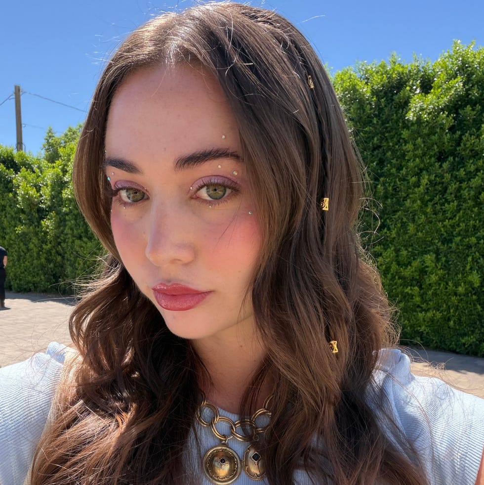 Outdoors, a person with long hair wearing a necklace is visible, background includes greenery and blue sky.