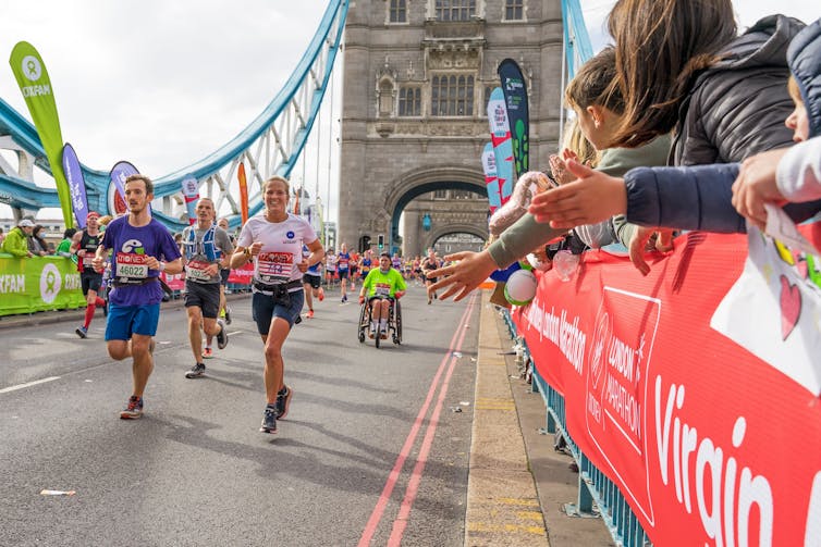 A group of marathon runners run down Tower Bridge, while spectators lean over a metal barrier to give them a high-five.
