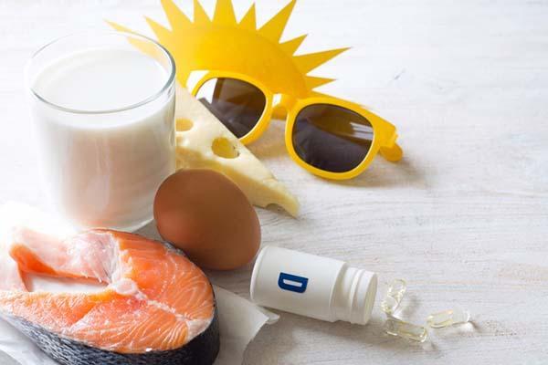 Arrangement of items representing ways to get vitamin D: yellow cardboard sun and yellow sunglasses, glass of milk, swiss cheese, egg, salmon steak, bottle of vitamin D supplement gel capsules with some spilled out.