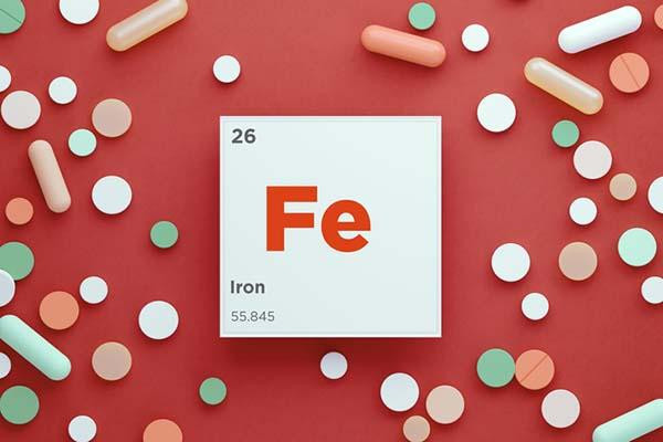 Pills in a variety of shapes and colors scattered on a red background surrounding a white square with the letters Fe, the chemical symbol for iron.