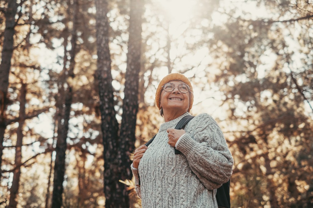 A happy older woman wearing a yellow beanie, glasses, and backpack, smiling and looking up at the sun shining through trees in a forest.
