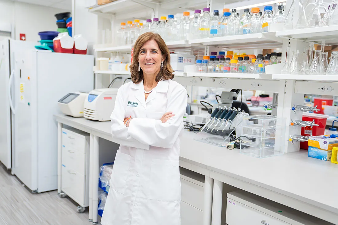 Dr. Pilar Alcaide in a white UHealth lab coat stands outdoors on the University of Miami campus near a fountain, holding both hands forward in a “U” hand gesture.