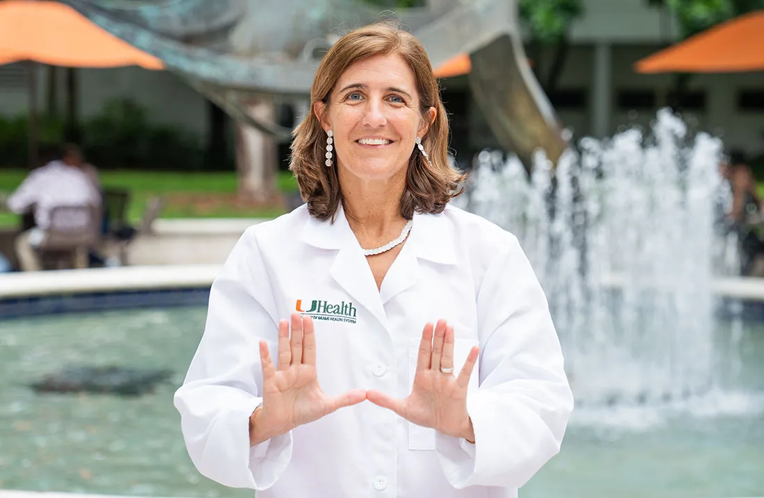 Dr. Pilar Alcaide in a white UHealth lab coat stands outdoors on the University of Miami campus near a fountain, holding both hands forward in a “U” hand gesture.