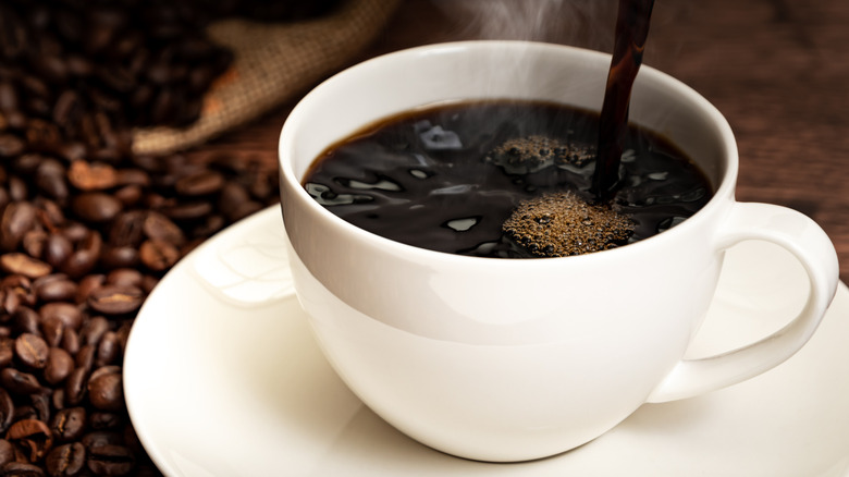cup of black coffee being poured into white cup on saucer next to some coffee beans