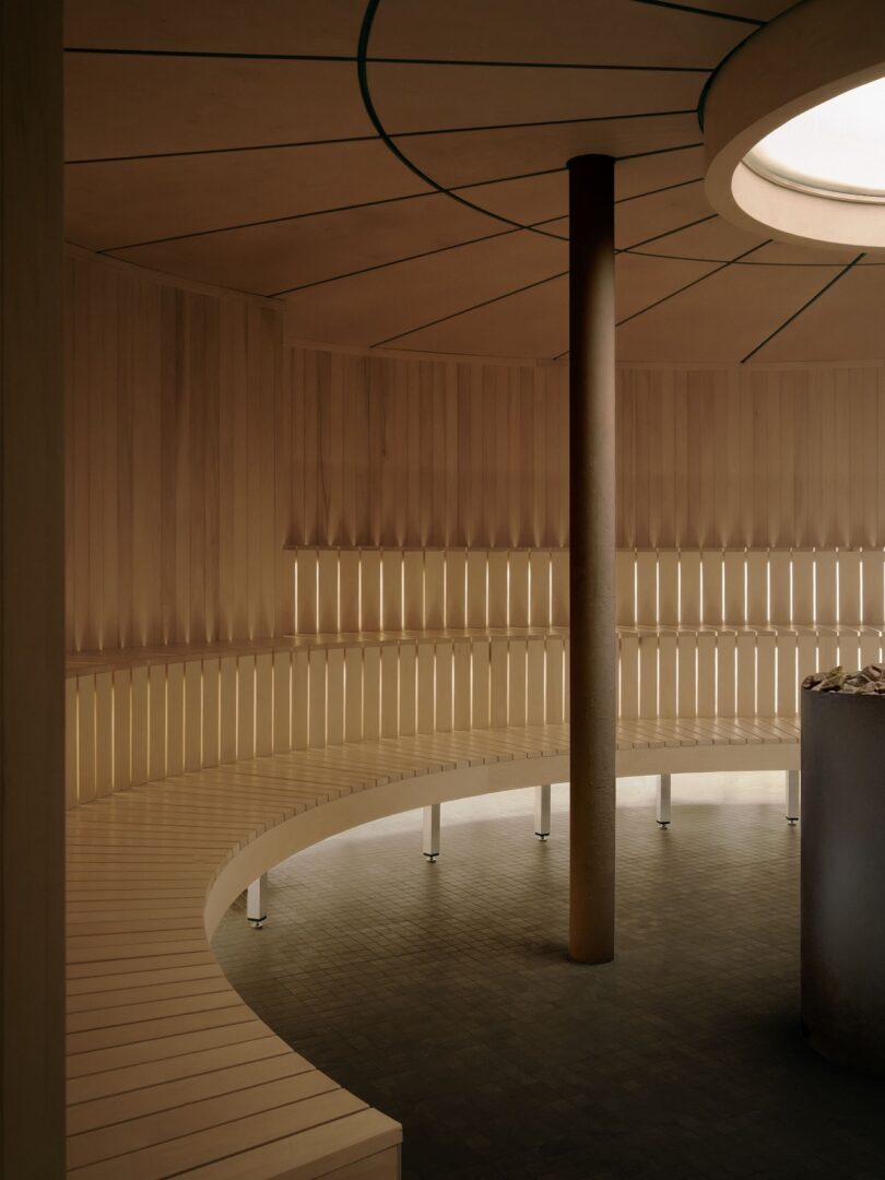 Circular wooden sauna interior with slatted walls, curved bench seating, central pillar, skylight, and a stone heater on a tiled floor.