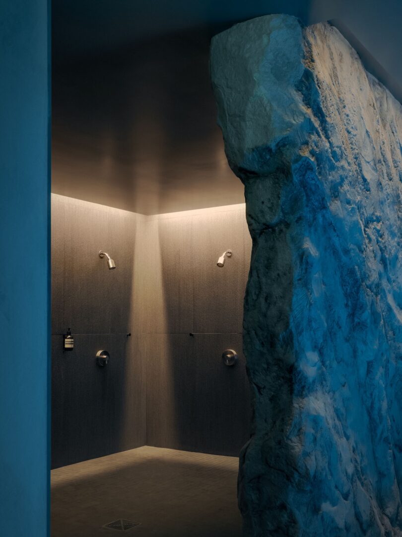 A modern shower area with two shower heads, gray tiled walls, and a large, textured stone wall partially lit with blue lighting.