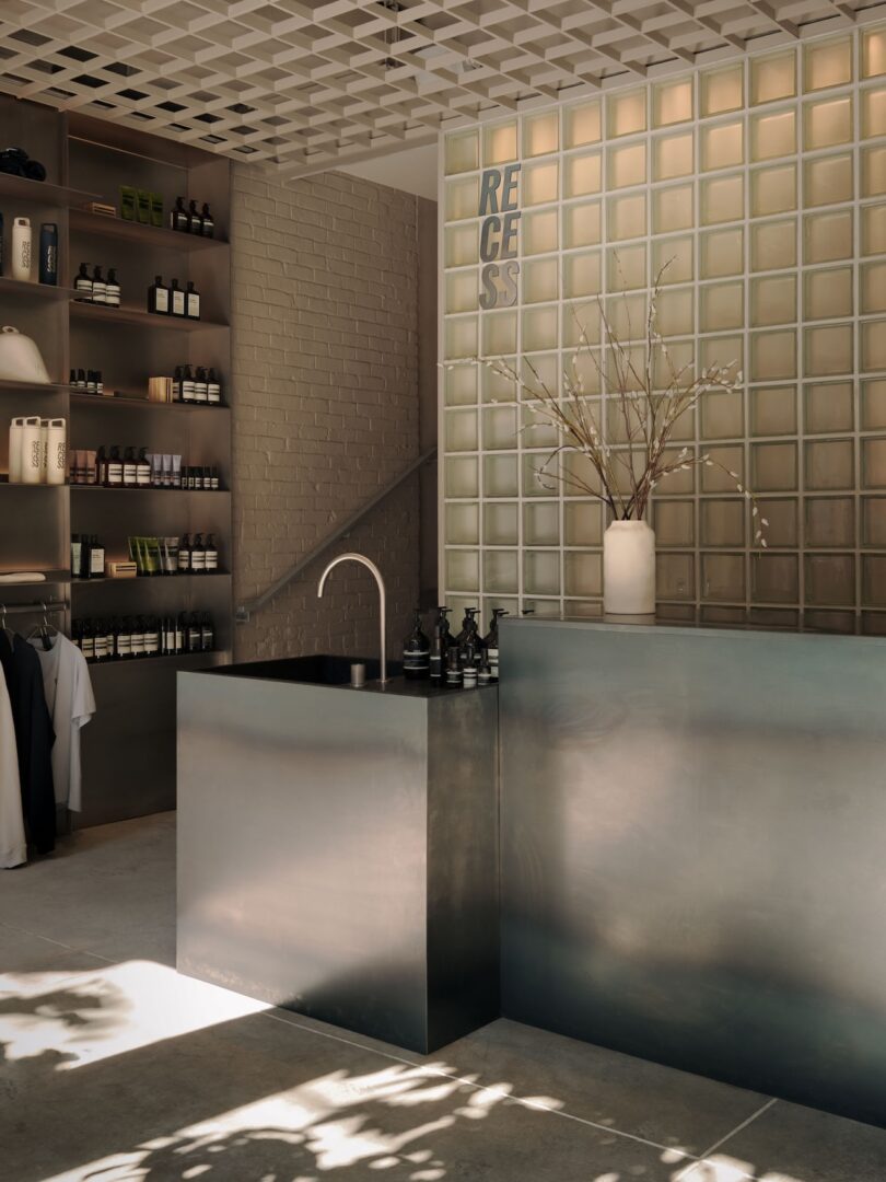 Minimalist retail store interior with metal counter, glass brick wall, shelves of products, a white vase with branches, and soft natural light.